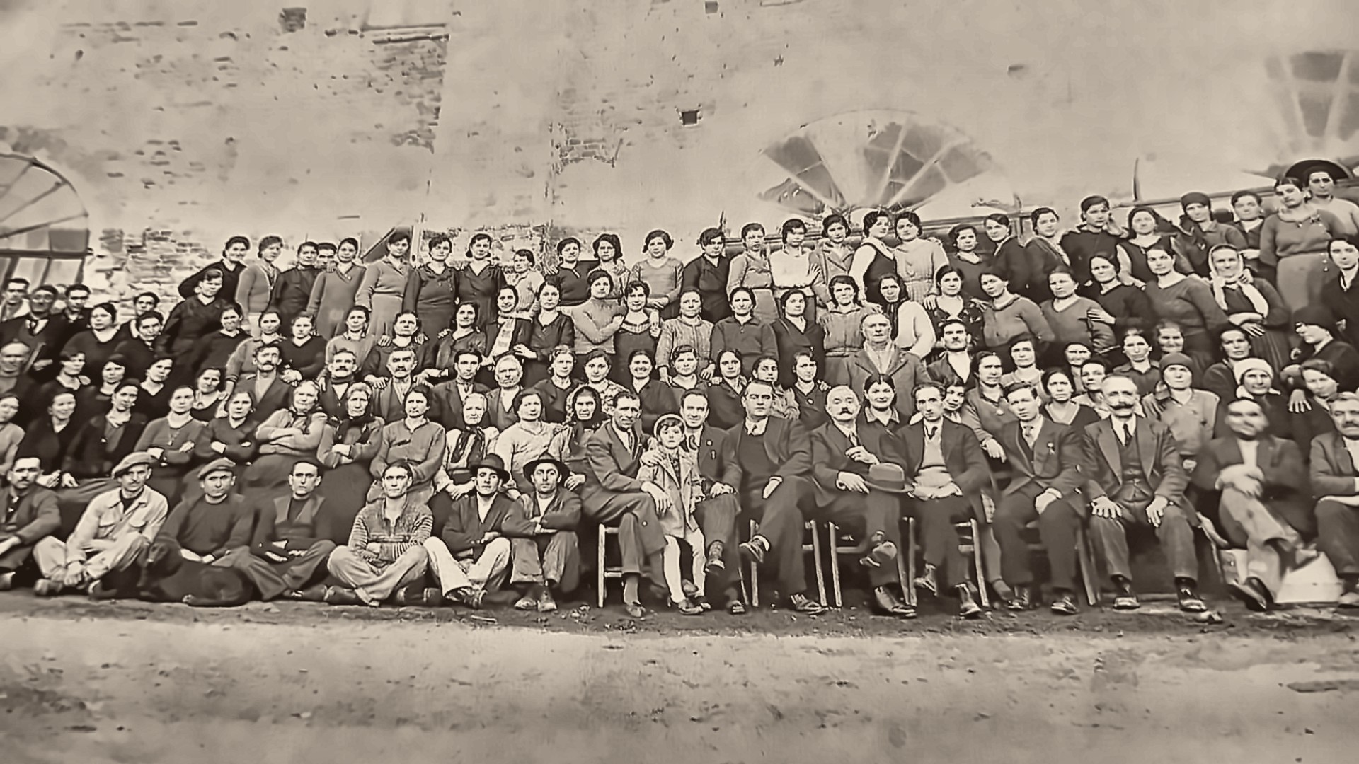 Fotografia di gruppo in bianco e nero, circa 1935: centinaia di operai e operaie in posa davanti a un muro di mattoni con finestre a ventaglio